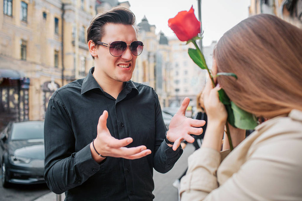 Young man stand outside in front of woman and talk to her. She holds red rose in hands. Guy wear sunglasses. - Photo, image