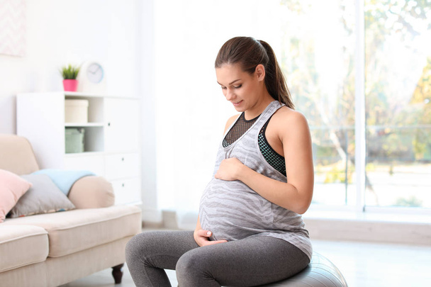 Young pregnant woman in fitness clothes sitting on exercise ball at home - Foto, imagen
