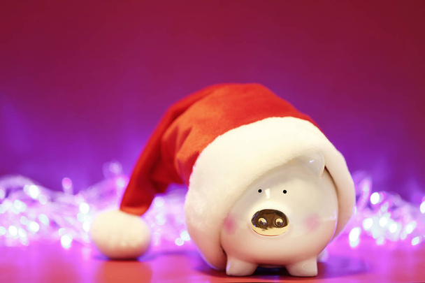 Piggy bank with Santa hat on table against blurred Christmas lights. Space for text - Valokuva, kuva