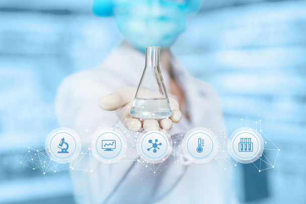 A laboratory assistant is showing a flask with transparent substance inside at the laboratory equipment icons foreground. The concept is the laboratorian researchers. - Photo, Image