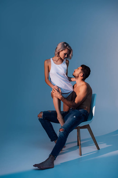 shirtless man sitting on chair and looking at beautiful woman on dark blue background - Фото, изображение