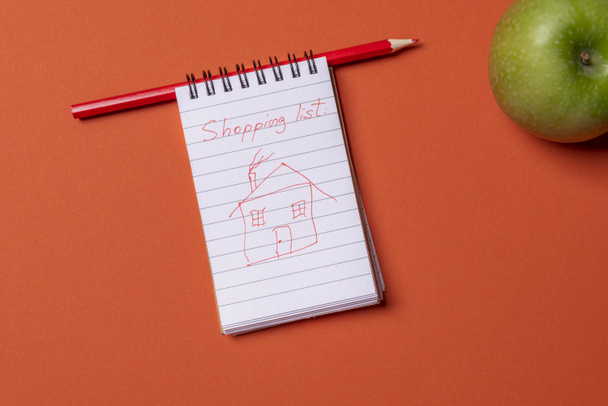 Σκίτσο σπίτι να σχεδιάσετε με ένα κόκκινο κραγιόν στη λίστα αγορών, σε μια σελίδα του σημειωματάριο σπιράλ. Παραπάνω άποψη με αντίγραφο χώρο σε ένα πορτοκαλί χρωματιστό παρασκήνιο - Φωτογραφία, εικόνα