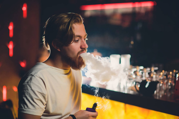 Young handsome hipster man with beard sitting in cafe with a cup of coffee, vaping and releases a cloud of vapor. - Foto, immagini