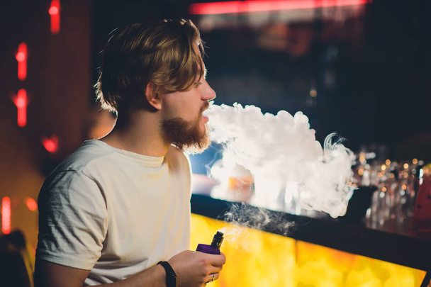 Young handsome hipster man with beard sitting in cafe with a cup of coffee, vaping and releases a cloud of vapor. - Foto, immagini