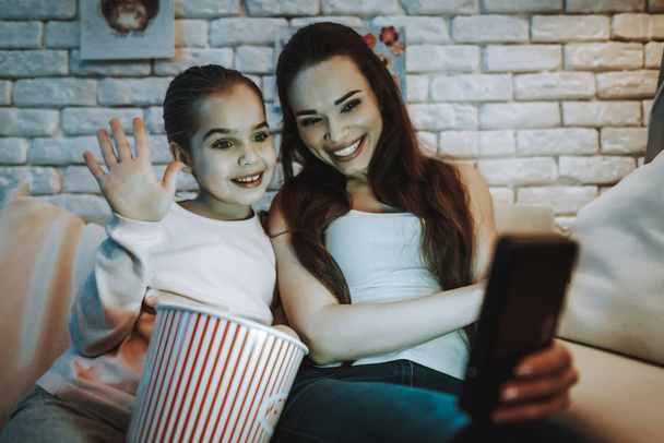 Mother with Daughter is Sitting on Couch. People Have a Video Call on Mobile Phone. Girl Eating a Popcorn. Girl is Waving. Persons is Smiling. Wall with Pictures. Evening Time. Home Interior. - Photo, image