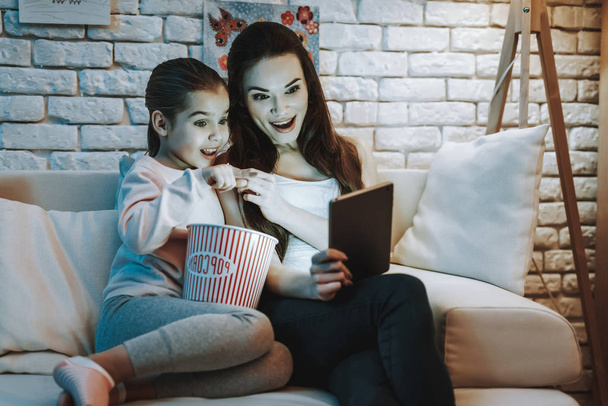 Mother with Daughter is Sitting on Couch. People Watching a Video on Laptop. Girl Eating a Popcorn. Girl Pointing on Screen. Persons is Smiling. Wall with Pictures. Evening Time. Home Interior. - Photo, image