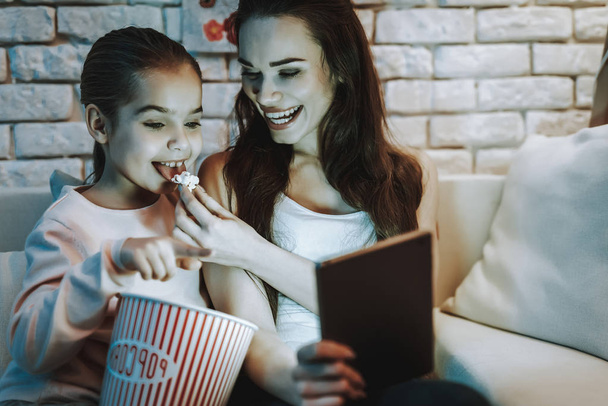 Mother with Daughter is Sitting on Couch. People Watching a Video on Laptop and Smiling. Mother Feeding a Daughter a Popcorn. Girl Pointing on Screen. Wall with Picture. Evening Time. Home Interior. - Zdjęcie, obraz