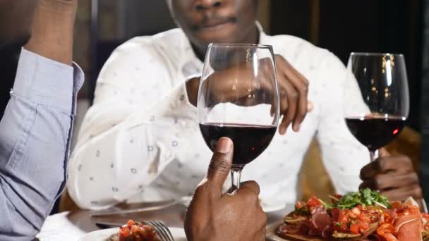 Twee unrecognizible Afrikaanse vrienden in restaurant over glas wijn aan tafel zit. Mannelijke Afrikaanse hand met een glas wijn. 4k - Video