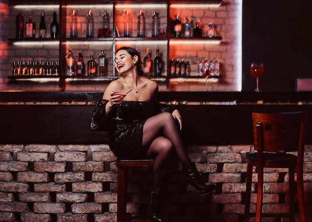 Une belle femme joyeuse portant une robe noire brillante assise à un comptoir de bar
 - Photo, image