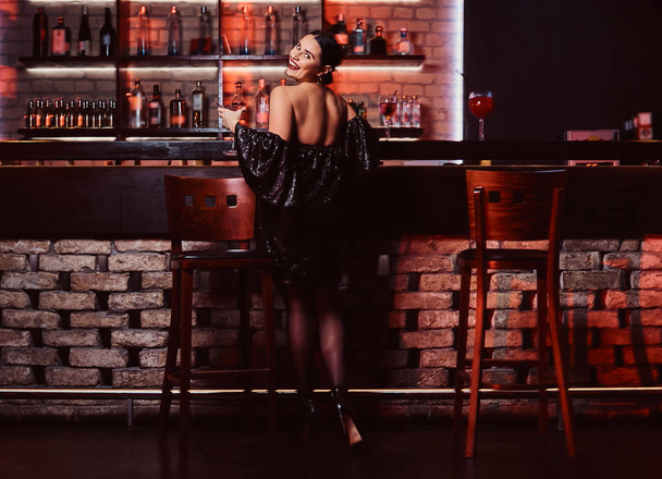 A seductive beautiful woman wearing a black shiny dress holding a cocktail while posing at a bar counter - Фото, зображення