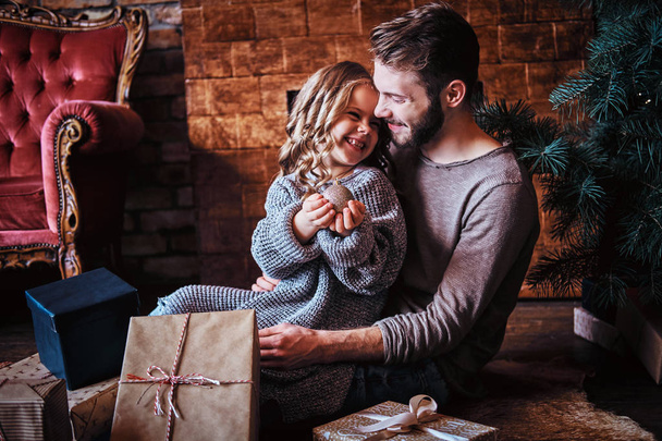 Glück niedliches kleines Mädchen hält Weihnachtskugel, während sie mit ihrem Vater auf einem Boden sitzt, der von Geschenken umgeben ist - Foto, Bild