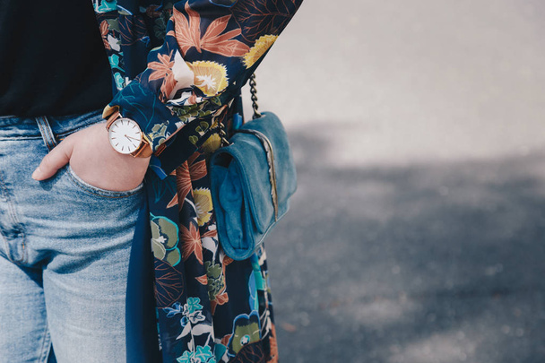 Modedetails im Streetstyle. Nahaufnahme, junge Modebloggerin in floraler Jacke und weiß-goldener analoger Armbanduhr. Zeitkontrolle, in der Hand eine schöne Wildledertasche. - Foto, Bild