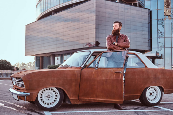 Stilvoller bärtiger Mann in altmodischer Kleidung lehnt auf einem getunten alten Retro-Auto mit offener Tür auf dem Stadtparkplatz in der Nähe eines Wolkenkratzers. - Foto, Bild