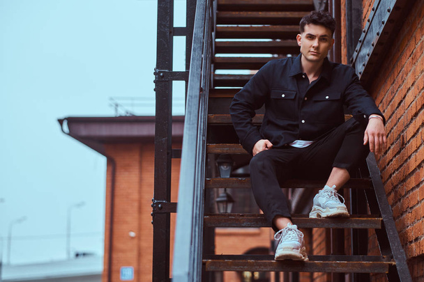 A fashionable guy sitting on the stairs outside the building with the industrial exterior. - Fotó, kép