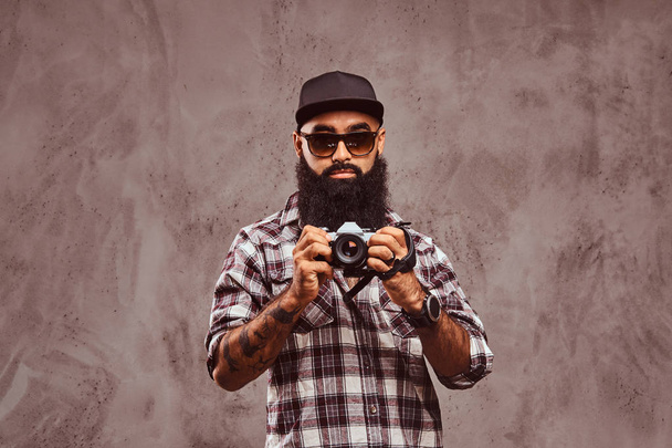 Yakın çekim kareli gömlek güneş gözlüğü ve bir kamera tutarak kap giyen sakallı bir Arap Erkek portresi.  - Fotoğraf, Görsel