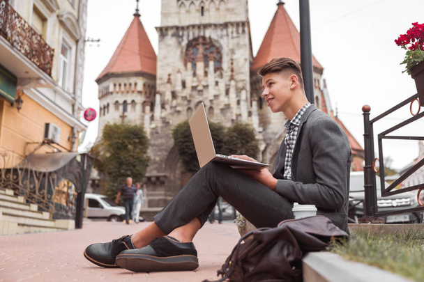 Side view of the handsome young man who is sitting outdoor against the background of old gothic build and using the laptop - Foto, Imagen