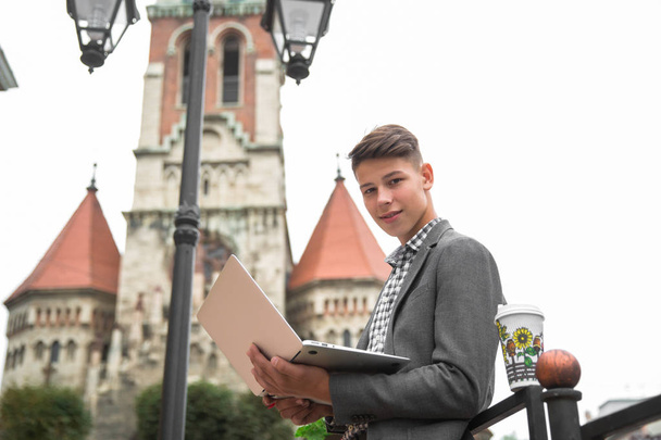 Side view of the smiling young man who is standing outdoor against the background of old gothic build and using the laptop - Foto, Bild