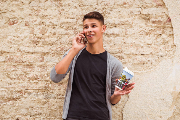 Portrait of the young man who is standing outdoor near the old house wall and speaking by phone and holding the coffee cup in hand - Foto, immagini