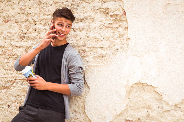 The young man standing outdoor near the old house wall and speaking by phone and holding the coffee cup in hand - Foto, immagini