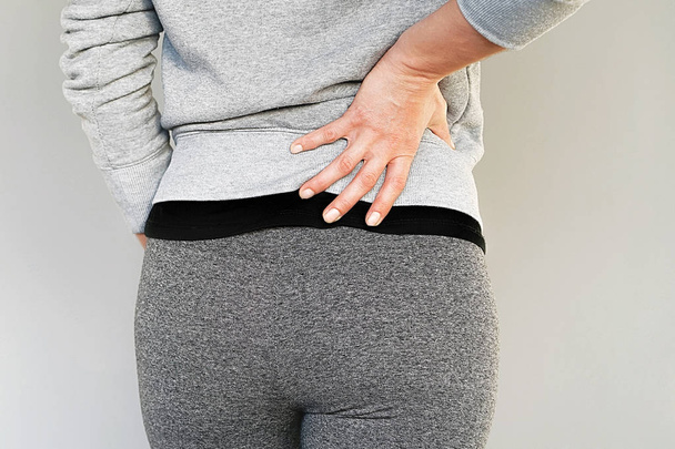 Young woman with back ache clasping her hand to her lower back. Close-up view of a young woman with pain in kidneys isolated on a gray background. Woman suffering from ribbing pain, waist pain. - Photo, Image