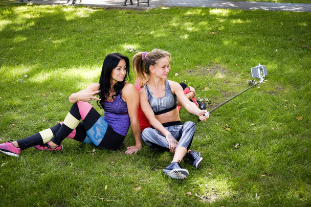Three sportive female friends sitting on a grass at the park outdoors, make selfie on a phone with a stick - Фото, изображение