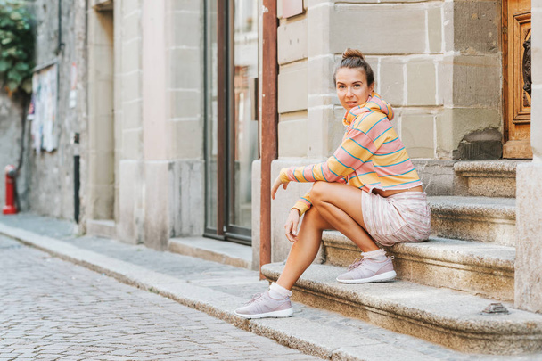 Outdoor portrait of young pretty woman sitting on steps in the city, wearing pink and yellow stripe sweatshirt, skirt, comfy purple sneakers - Photo, Image