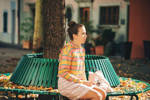 Herfst portret van jonge mooie vrouw zittend op het bankje in zonlicht, stripe sweatshirt en mini rok dragen - Foto, afbeelding