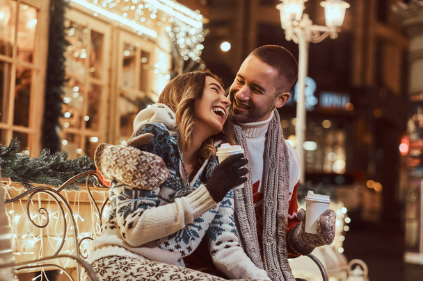 A young romantic couple wearing warm clothes outdoor at Christmas time, sitting on a bench and warming with hot coffee in evening street decorated with beautiful lights. - Фото, изображение