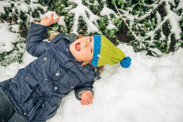 fröhlicher kleiner Junge spielt draußen mit Schnee, trägt warme Winterjacke und Strickmütze, liegt im Schnee, von oben gesehen - Foto, Bild
