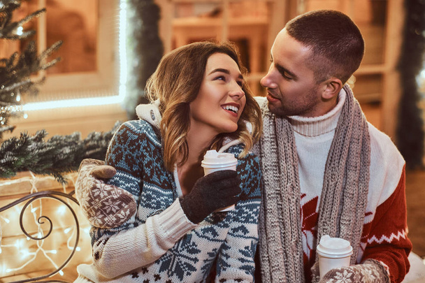 A young romantic couple wearing warm clothes outdoor at Christmas time, sitting on a bench and warming with hot coffee in evening street decorated with beautiful lights. - Photo, Image