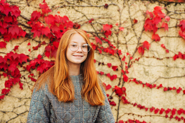 Autumn portrait of adorable red-haired preteen girl girl check jacket and glasses, posing next to wall with red ivy leaves, fall fashion for kids - Photo, Image