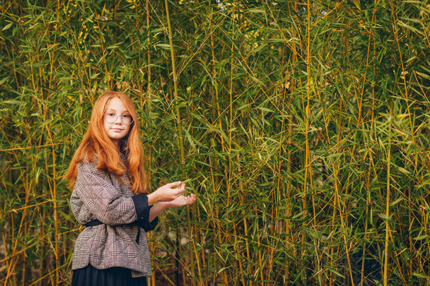 Retrato de cerca al aire libre de una hermosa niña preadolescente pelirroja con abrigo de cheques y anteojos. Posando en fondo de bambú
 - Foto, Imagen