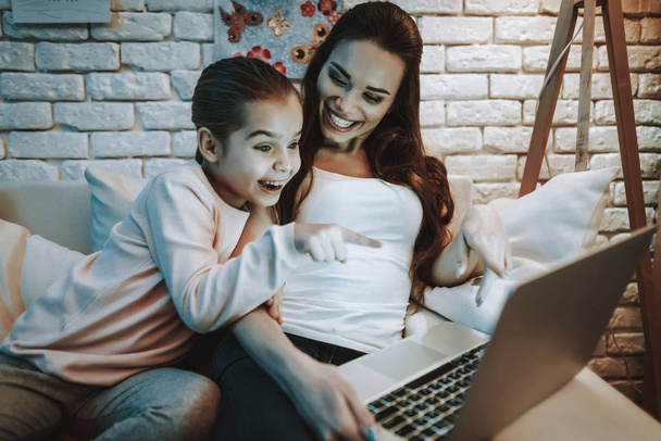 Mother with Daughter is Sitting on Couch. People Watching a Video on Laptop. Girl Pointing on Screen to Mother. People is Smiling. Wall with Pictures on Background. Evening Time. Home Interior. - Fotografie, Obrázek