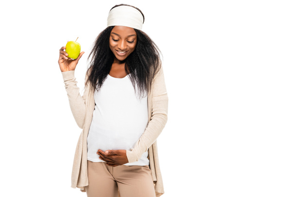 africká americká těhotná žena v ruce drží jablko a při pohledu dolů izolované na bílém - Fotografie, Obrázek