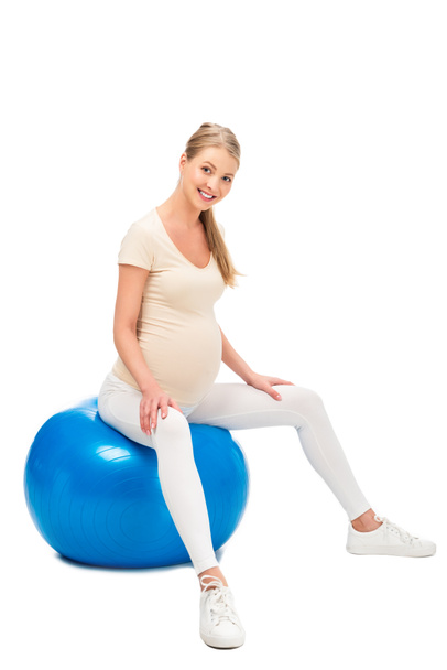 mujer rubia embarazada sentada en la pelota de fitness y cogidas de las manos en las rodillas aisladas en blanco
 - Foto, imagen