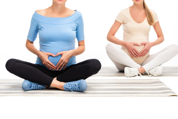 abgeschnittene Ansicht von zwei schwangeren Frauen mit Händen auf Bäuchen in Herzzeichen isoliert auf weiß - Foto, Bild