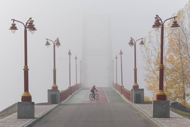 Женщина в красной куртке катается на велосипеде по мосту туманным утром, Киев, Украина
. - Фото, изображение