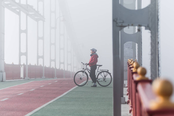 Woman in red jacket riding bicycle on a bridge in foggy morning, Kyiv, Ukraine. - 写真・画像