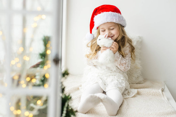 kleines Mädchen in weißem Kleid mit einem kleinen Mädchen in rotem Kleid, das am Fenster sitzt und auf Weihnachten und Weihnachtsmann wartet - Foto, Bild