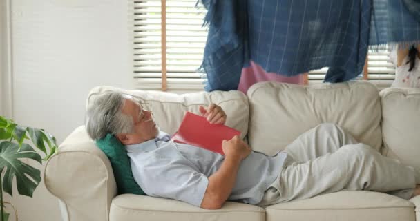 Senior man sleeping with book on the sofa in living room and his wife put blanket on him. - Metraje, vídeo
