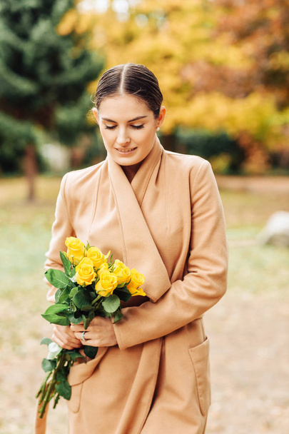 Outdoor portrait of beautiful young woman wearing camel wrap coat, holding yellow roses bouquet - Valokuva, kuva