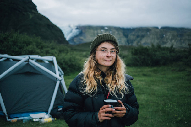 Young beautiful blonde woman holding coffee mug and standing outdoors in mountains at camp tent  - Photo, image