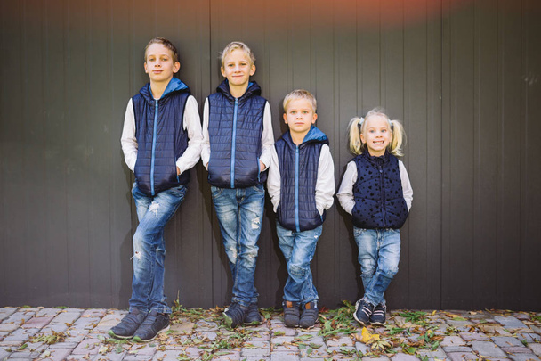 Funny Caucasian big family of three brothers and sister posing standing on growth background of wall in full growth. Equally stylishly dressed in blue vests and jeans. Theme girl in male circle. - Foto, immagini
