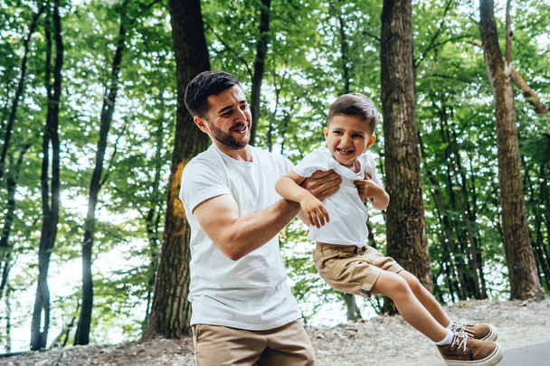Onnellinen nuori isä kiertelee poikansa sylissä kesäpuistossa.
. - Valokuva, kuva
