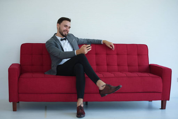 Nuori mies istuu ja käyttää älypuhelinta punaisella sohvalla
. - Valokuva, kuva