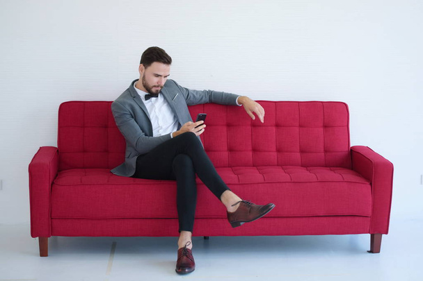 Un jeune homme assis et utilisant un téléphone intelligent sur un canapé rouge
. - Photo, image