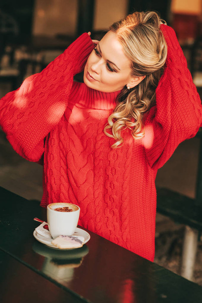 Portrait of beautiful elegant woman with cup of cappuccino in outdoor cafe, wearing red pullover, fixing hair - Foto, immagini
