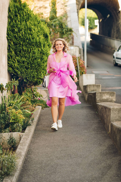 Outdoor portrait of beautiful elegant woman walking down the street, wearing pink belted dress - Photo, image