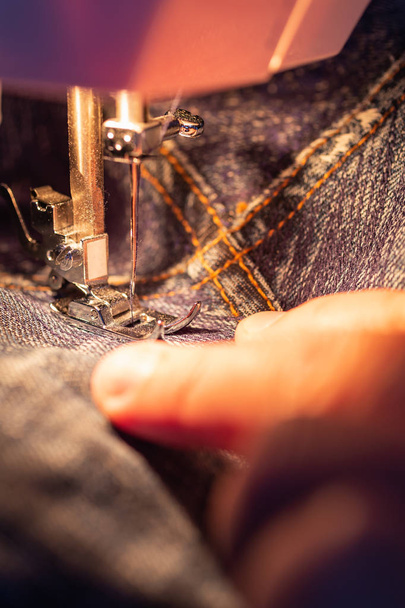 Reparação de jeans na máquina de costura. Vista do tecido, agulha e linha. Iluminação da lâmpada incandescente embutida. Jeans são um tipo de calças, tipicamente feitas de ganga ou pano de calça
 - Foto, Imagem