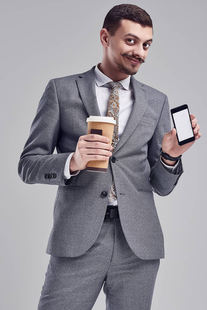 ファッション灰色完全なスーツに派手な口髭のハンサムな若い自信アラビア語ビジネスマンの肖像一杯のコーヒー、スタジオの背景に携帯電話を保持しています。 - 写真・画像